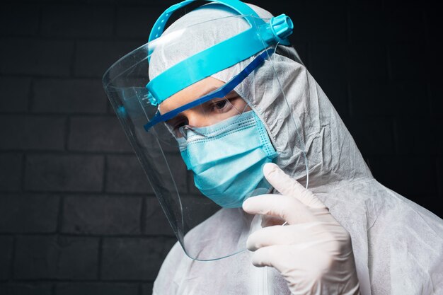 Portrait côté studio d'un jeune médecin portant une combinaison EPI contre le coronavirus et le covid-19. mur de mur de briques noires.