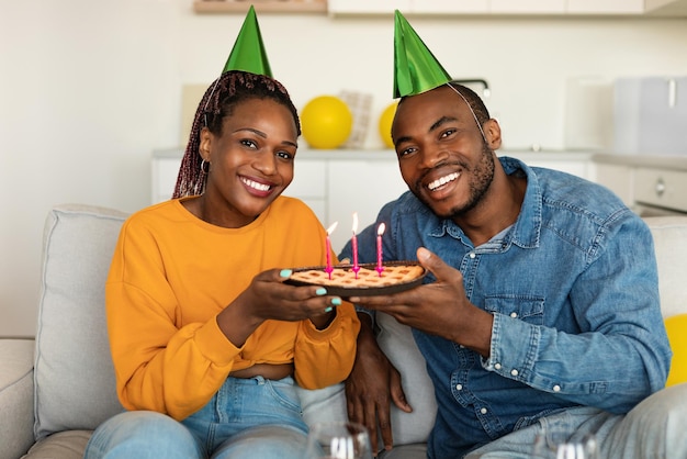 Portrait d'un couple afro-américain en chapeaux de fête tenant une tarte d'anniversaire avec des bougies allumées et souriant à la caméra à la maison