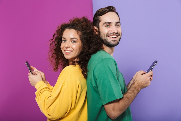 Portrait d'un couple caucasien positif dans des vêtements colorés souriant et tenant des téléphones portables debout dos à dos isolés