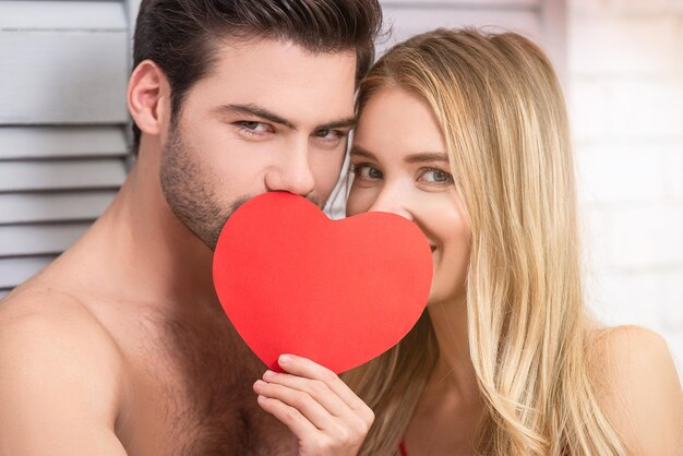 Portrait de couple couvrant les visages avec du coeur de papier rouge à la maison