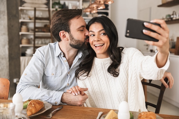 Portrait d'un couple européen heureux homme et femme prenant une photo de selfie tout en prenant son petit-déjeuner dans la cuisine à la maison