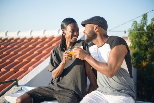 Portrait d'un couple gay africain exprimant l'amour. Deux hommes barbus datant sur le toit assis près souriant tenant des verres de jus en se regardant avec tendre. Concept d'amour et de vie des couples LGBT