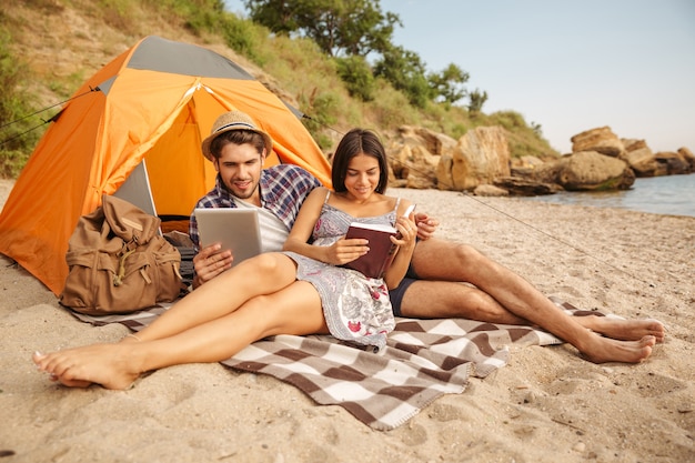 Portrait d'un couple heureux assis à la tente à l'aide d'une tablette et d'un livre de lecture