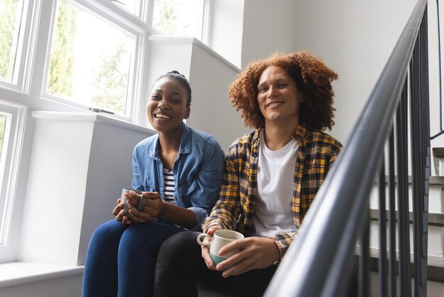 Photo portrait d'un couple heureux et diversifié assis sur les escaliers à la maison en train de boire du café. la convivialité, la relation, le temps libre, le style de vie et la vie domestique, inchangés.