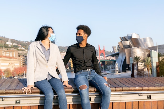 Portrait d'un couple de jeunes amis assis à Bilbao avec un ciel bleu heureux avec des masques faciaux en raison de la pandémie de coronavirus Covid-19 2020