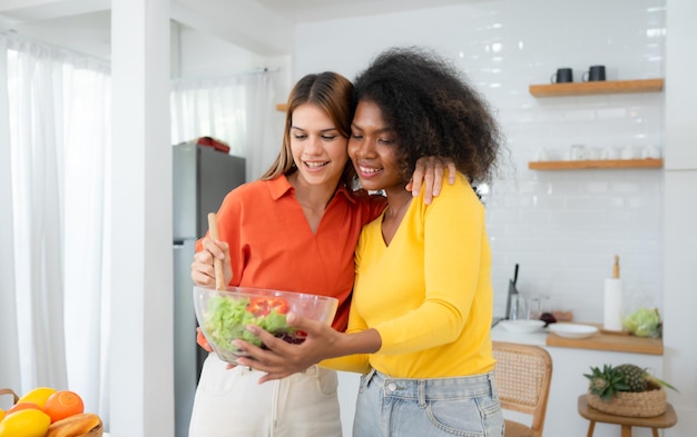 Portrait d'un couple de jeunes femmes LGBT mangeant de la salade et souriant alors qu'ils se tiennent dans la cuisine à la maison