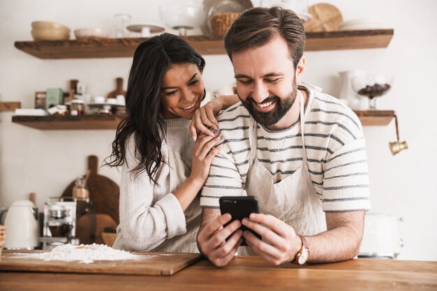 Portrait d'un couple joyeux homme et femme de 30 ans portant des tabliers lisant une recette pendant la cuisson de la pâtisserie avec de la farine et des œufs dans la cuisine à la maison