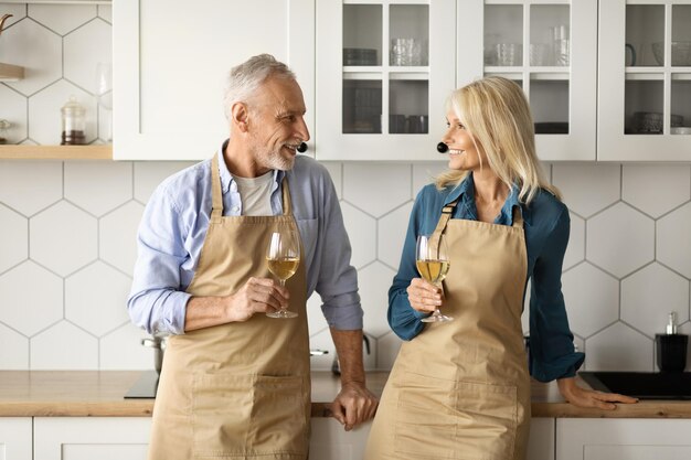 Portrait d'un couple de personnes âgées romantique en tabliers buvant du vin ensemble dans la cuisine