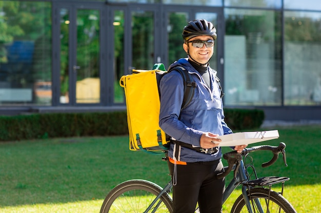 Portrait de courrier de vélo avec sac jaune et vélo. Homme, casque, lunettes, tenue, boîte pizza