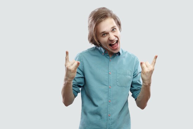 Portrait de crier beau jeune homme blond aux cheveux longs en chemise bleue décontractée debout avec signe rock et regardant la caméra en criant. tourné en studio intérieur, isolé sur fond gris clair.