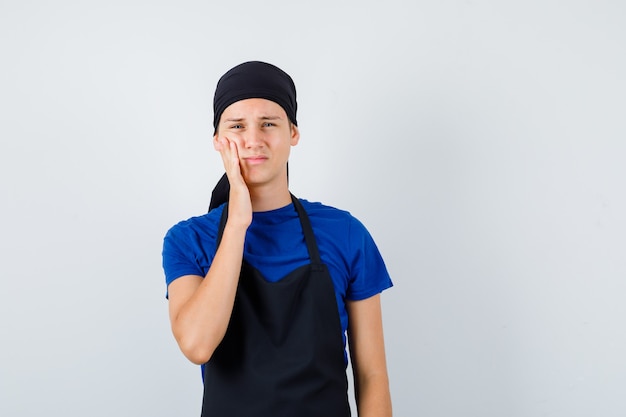 Portrait d'un cuisinier adolescent souffrant de maux de dents en t-shirt, tablier et à la vue de face douloureuse