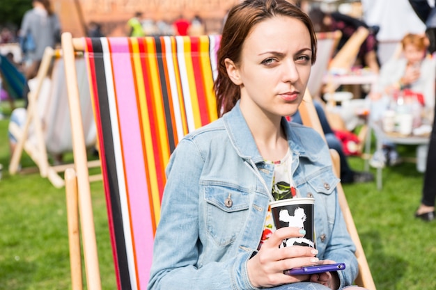 Portrait décontracté d'une jeune femme brune portant une veste en jean et tenant un téléphone portable et une tasse de café à emporter tout en étant assise sur une chaise de plage rayée dans le champ au pique-nique ou au festival en plein air