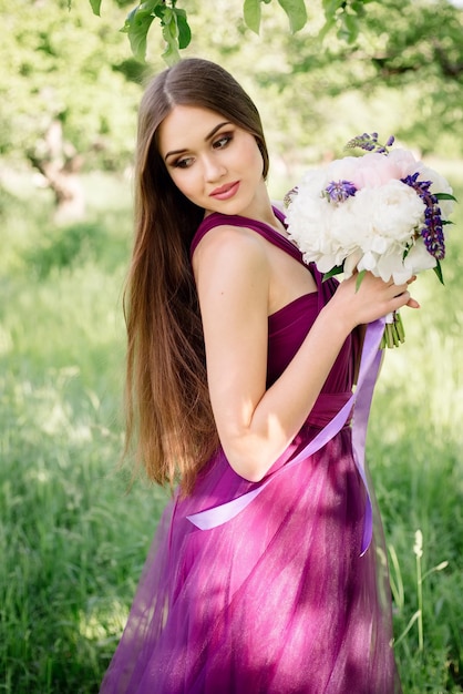 Portrait de demoiselle d'honneur bouquet de mariage luxueux de pivoines et de fleurs debout lors d'une cérémonie dans le jardin en robe violette violette souriant et regardant la caméra Journée ensoleillée diplôme d'études secondaires