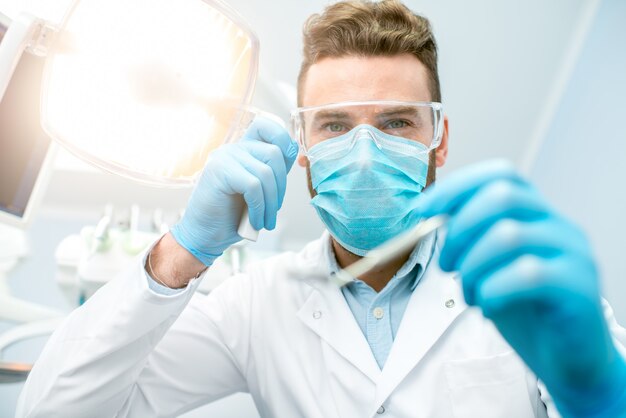 Portrait d'un dentiste effrayant en masque et lunettes de protection avec des outils dentaires pendant l'opération en regardant la caméra