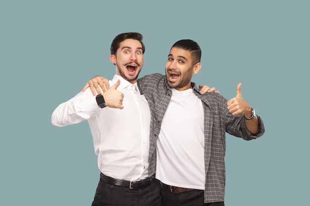 Portrait de deux beaux amis ou partenaires heureux barbus se serrant dans leurs bras et regardant la caméra avec un visage étonné satisfait et les pouces vers le haut. tourné en studio intérieur, isolé sur fond bleu clair.