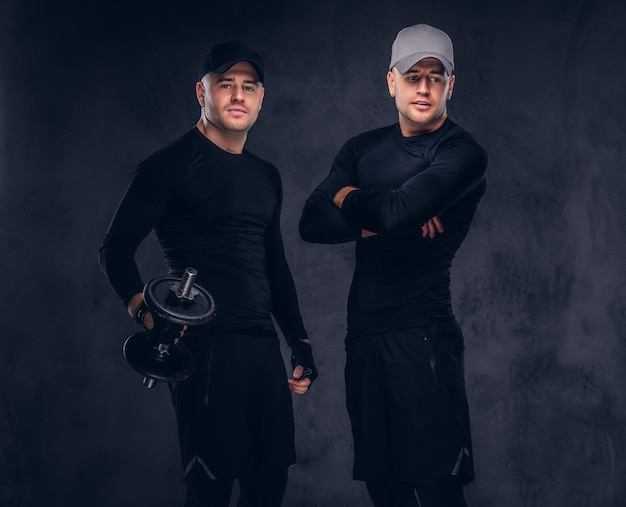 Portrait de deux beaux jeunes hommes vêtus d'un vêtement de sport noir et d'une casquette de baseball sur fond sombre, tient un haltère.