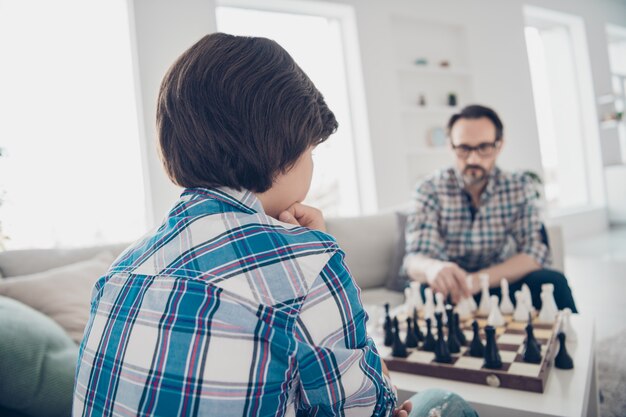 Portrait de deux beaux mecs sérieux concentrés qualifiés papa et fils pré-adolescent assis sur un canapé jouant aux échecs en mouvement de pièces en intérieur moderne blanc clair salon-salle