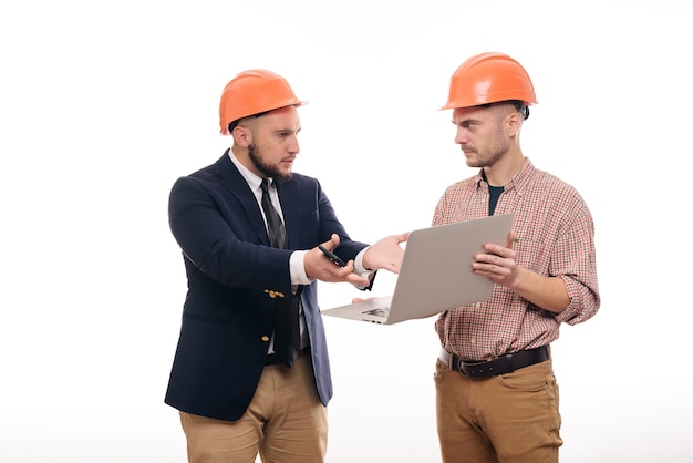 Portrait de deux constructeurs dans des casques de protection orange debout sur fond blanc isolé et regardant l'écran de l'ordinateur portable. Discuter du projet de construction