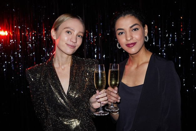 Portrait de deux élégantes jeunes femmes tenant des verres à champagne et souriant à la caméra tout en posant sur fond mousseux à la fête, tourné avec flash