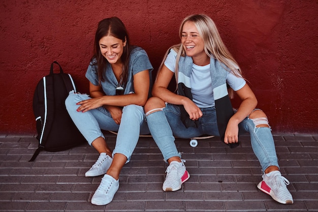 Portrait de deux filles hipster riantes assises ensemble sur une planche à roulettes et s'appuyant sur un mur sur le trottoir.