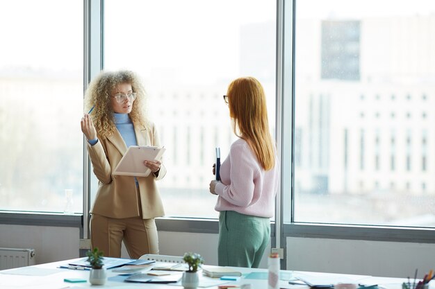 Portrait de deux jeunes femmes d'affaires discutant du projet de travail en se tenant debout contre la fenêtre dans un bureau moderne, copiez l'espace