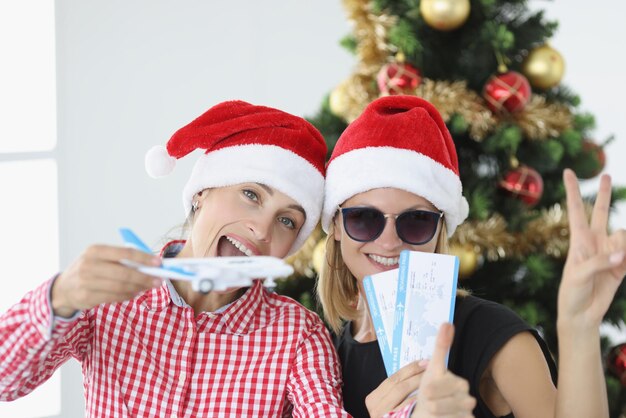 Portrait de deux jeunes femmes en chapeaux de père Noël tenant un avion et des billets contre de nouveaux