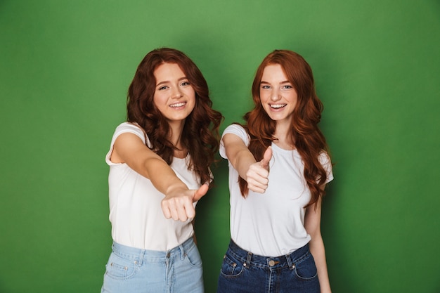 Portrait de deux jeunes femmes rousses de 20 ans en tenue décontractée souriant à la caméra et montrant les pouces vers le haut, isolé sur fond vert