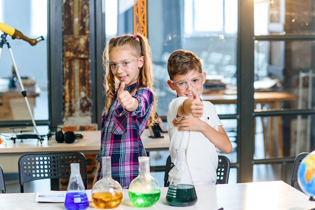 Portrait de deux jeunes scientifiques gais de race blanche dans des lunettes de sécurité montrant les pouces vers le haut en classe chimique de l'école primaire