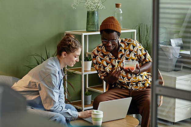 Portrait de deux jeunes utilisant un ordinateur portable tout en se relaxant dans un salon de bureau derrière un mur de verre