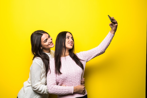 Portrait de deux jolies filles vêtues de chandails debout et prenant un selfie isolé sur mur jaune