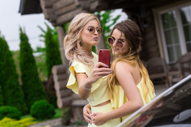 Portrait De Deux Jolies Jeunes Demoiselles D'honneur En Robes Jaunes à Proximité D'une Maison En Bois à La Mode. Concept De Mode De Vie Des Gens. Faire Selfie Shot Sur Téléphone Mobile En Souriant Et En Envoyant Un Baiser Aérien