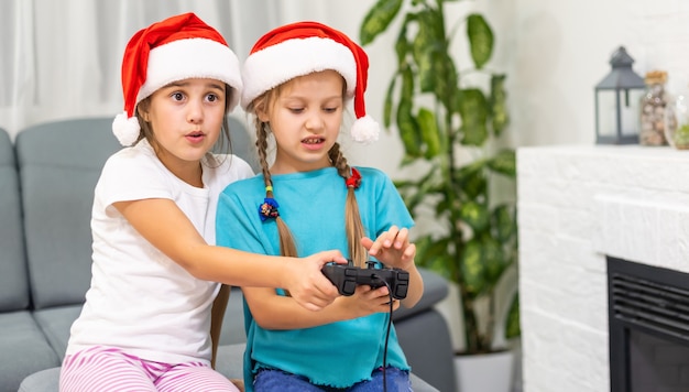 Portrait de deux jolies jolies jolies jolies jolies charmantes filles positives gaies drôles et gaies en chapeaux de père Noël assis sur un divan jouant à la bataille d'un appareil vidéo