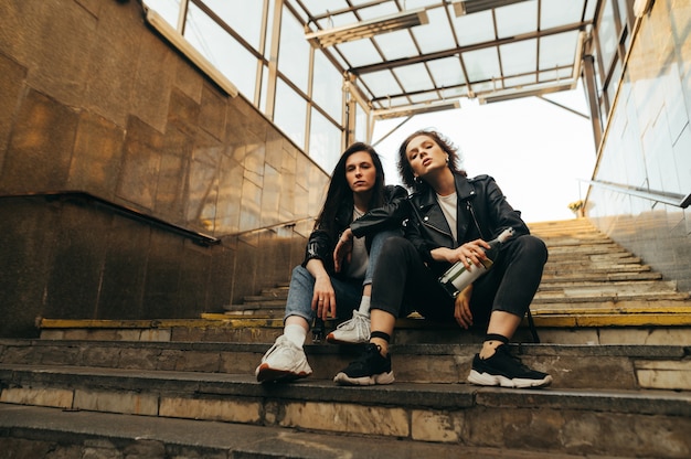 Portrait de deux modèles en vêtements décontractés élégants assis sur les escaliers avec une bouteille de vin à la main