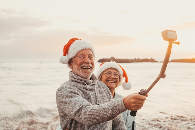 Portrait de deux personnes âgées mignonnes s'amusant et s'amusant ensemble à la plage les jours de noël à la plage portant des chapeaux de noël regardant et tenant une caméra prenant des vidéos de vacances
