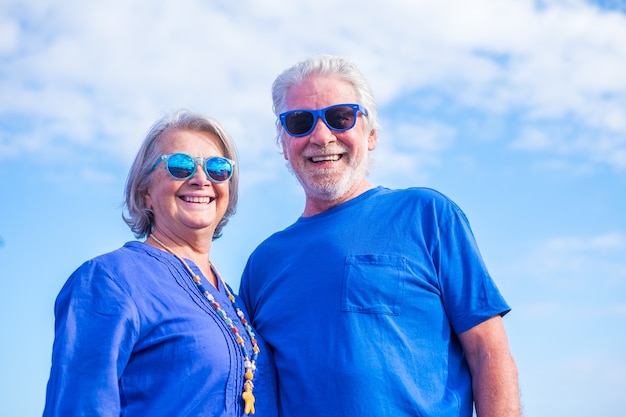 Portrait de deux personnes mûres souriant et regardant la caméra portant des vêtements bleus et des lunettes de soleil avec un ciel bleu en arrière-plan - des personnes âgées heureuses s'amusant ensemble