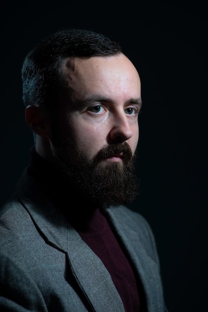 Un portrait dramatique d'un homme du millénaire barbu dans une veste et un pull sur fond sombre