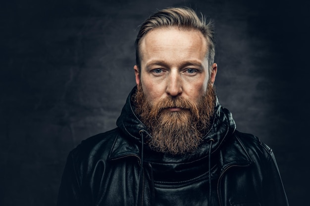 Portrait dramatique en studio d'un homme barbu roux vêtu d'une veste en cuir sur fond de vignette gris foncé.