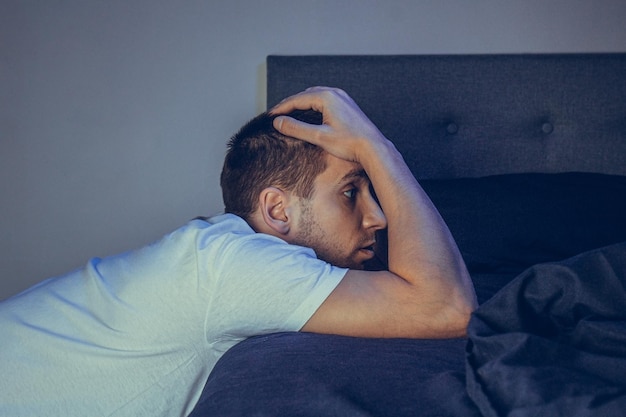 Portrait dramatique de style de vie d'un beau gars dans la trentaine et la quarantaine assis tristement sur le lit se sentant anxieux et souffrant de dépression homme attrayant déprimé et contrarié dans la chambre à coucher de la maison