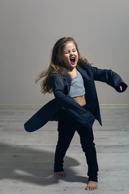 Le portrait drôle d'une fille à la mode dans une grande veste grise se dresse contre un mur gris à côté de la fenêtre de la maison