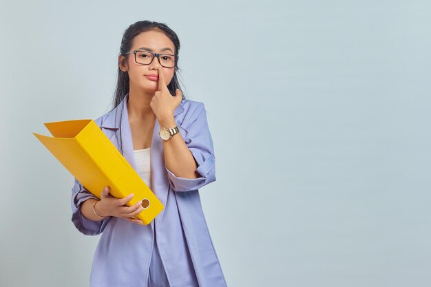 Portrait d'une drôle de jeune femme asiatique pensant à quelque chose tout en portant un dossier jaune et en choisissant son nez isolé sur fond jaune