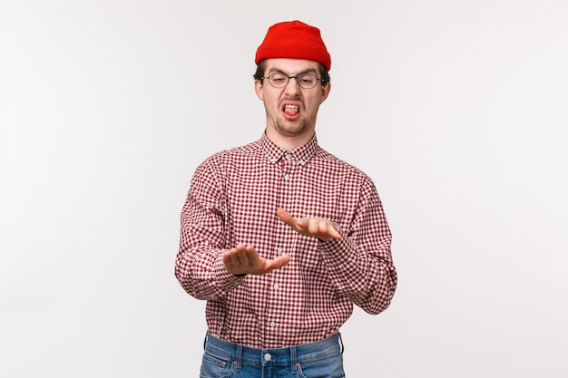 Portrait drôle jeune homme réticent en bonnet rouge et lunettes, se serrant la main dans le rejet, bâton de langue et grimaçant dégoûté, exprime sa désapprobation et n'aime pas ou horrible chose laide,