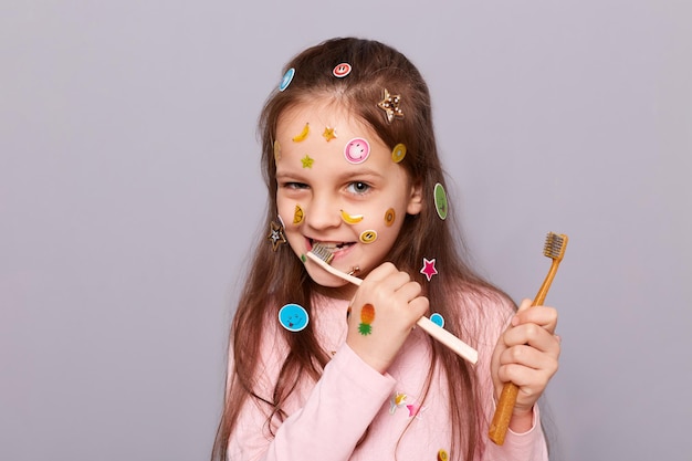 Portrait d'une drôle de petite fille ludique couverte d'autocollants colorés portant une chemise rose debout isolée sur fond gris se brossant les dents en prenant soin de sa bouche seule problèmes dentaires