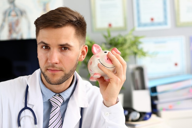 Portrait du docteur secouant la tirelire. Médecin vêtu d'un uniforme blanc avec stéthoscope et posant dans le bureau de l'hôpital. Homme regardant la caméra avec calme. Concept de police d'assurance maladie