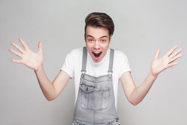 Portrait du gagnant jeune homme brune en t-shirt blanc décontracté et salopette en jean debout, bras levés et regardant la caméra avec un visage excité étonné. tourné en studio intérieur, isolé sur fond gris.