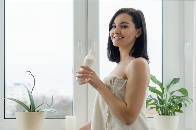 Portrait du matin de la jeune femme souriante dans une serviette de bain, buvant du lait