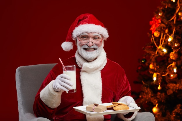 Portrait du père Noël assis sur un fauteuil avec du lait et des biscuits en souriant