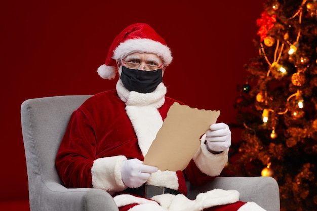 Portrait du père Noël portant un masque de protection en lisant une lettre alors qu'il était assis sur un fauteuil