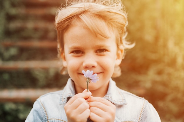 Portrait du visage d'un mignon heureux souriant petit garçon de cinq ans échevelé long blond aux yeux sombres caucasien enfant d'âge préscolaire garçon tenant une délicate fleur sauvage lilas pâle dans ses mains sur la nature flare