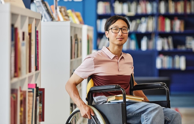 Portrait dynamique d'un jeune homme asiatique en fauteuil roulant regardant la caméra dans la bibliothèque du collège et souriant