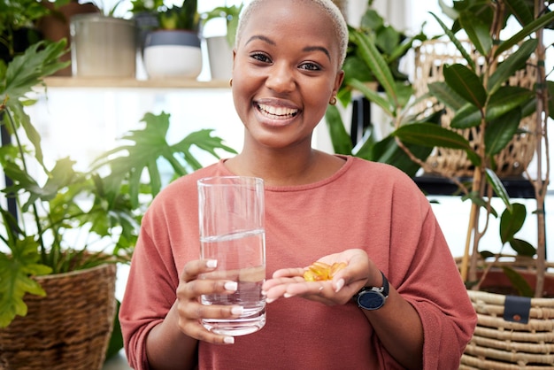 Portrait d'eau et de médicaments avec une femme noire dans sa maison tenant des vitamines ou des suppléments pour la santé Sourire en verre et en pilules avec une jeune femme heureuse buvant pour le bien-être ou l'hydratation